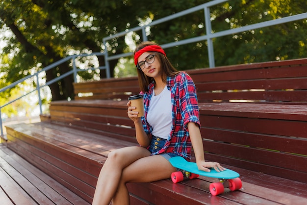 Young woman skater dressed in stylish clothes sits on the stands in a skatepark and drinks coffee on a bright sunny day