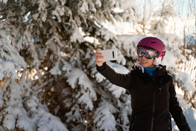 A young woman in skiing clothes, a pair of ski goggles and a ski helmet making a selfie in a ski resort next to a snowy tree