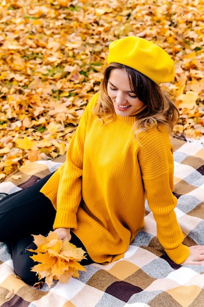 A young woman smiles looks at a bouquet of autumn maple leaves and sits on a blanket in the park