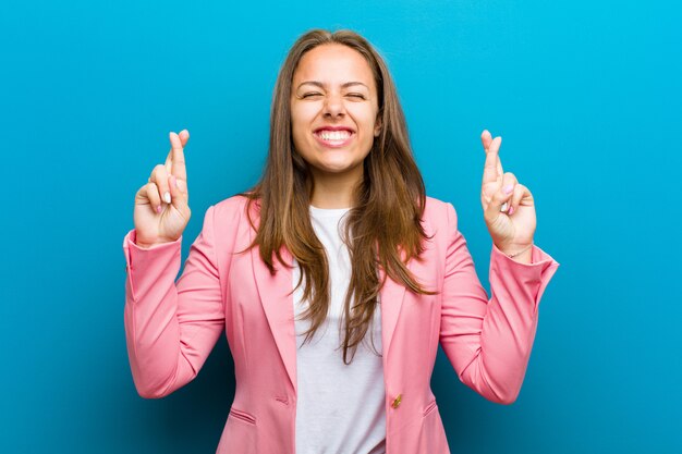 Young woman smiling and anxiously crossing both fingers, feeling worried and wishing or hoping for good luck against blue wall