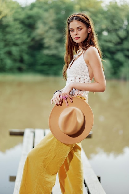 A young woman smiling in an image of a hippie and ecodress dancing in nature by the lake wearing a hat and yellow pants in the summer sunset