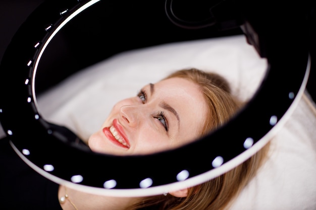 Young woman smiling portrait with pink pigment permanent tattoo lips laying under circle lamp