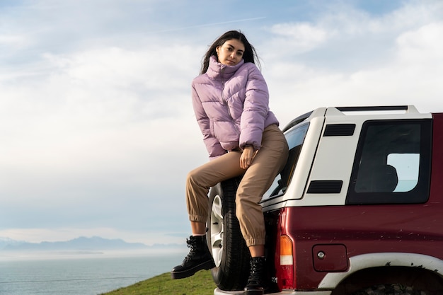 Young woman smiling seated on the rear wheel of an off road car with a beautiful seascape