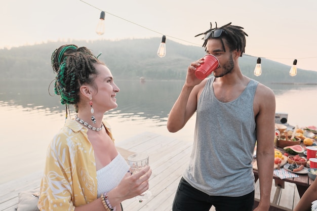 Young woman smiling to young man while they standing on a pier outdoors and drinking cocktails