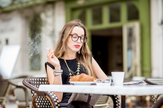 Young woman smoking a cigarette while having a breakfast outdoors at the typical french cafe terrace in France