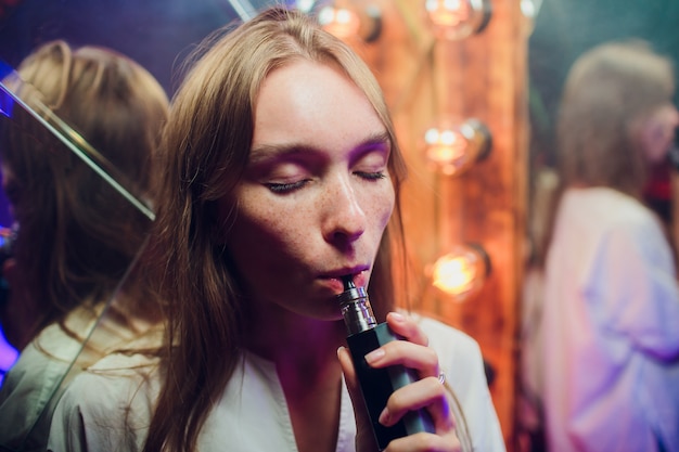 Young woman smoking electronic cigarette against background of mirrors