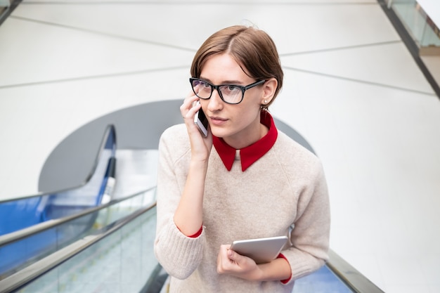 Young woman speaking on the phone at an escalator staircase. Female business person on a moving staircase using modern technology