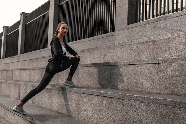 Young woman in sportive clothes is doing warming up exercises near the stadium. Legs stretching on stone stairs