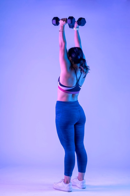 Young woman in sportswear from behind doing shoulder press with two dumbbells