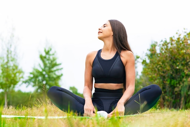 Young woman in sportswear meditates in yoga lotus position while sitting outdoors in a park in summe