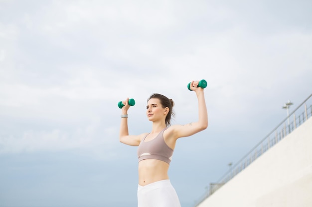 Young woman in sporty top doing exercises with dumbbells in hands thoughtfully looking aside while spending time outdoor