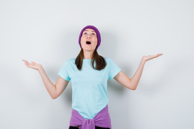 Young woman spreading hands in questioning manner, keeping mouth wide open in blue t-shirt, purple beanie and looking shocked , front view.