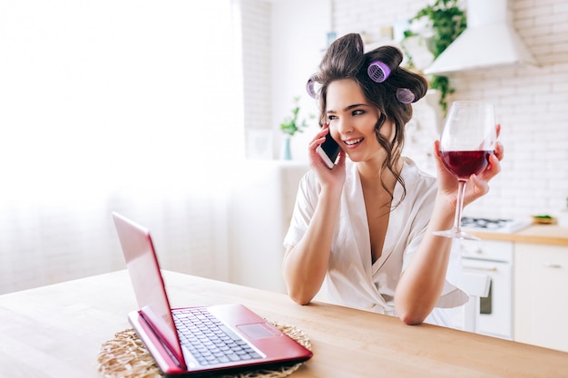 Young woman stand in kitchen and talking on phone. Holding red wine glass in hand. Housekeeper with curlers in hair. Housewife careless life.