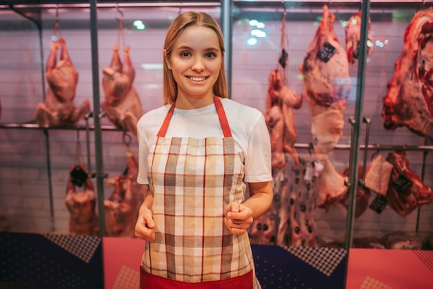 Young woman stand and meat shelf in grocery store. She pose on camera and smile. Positive female worker