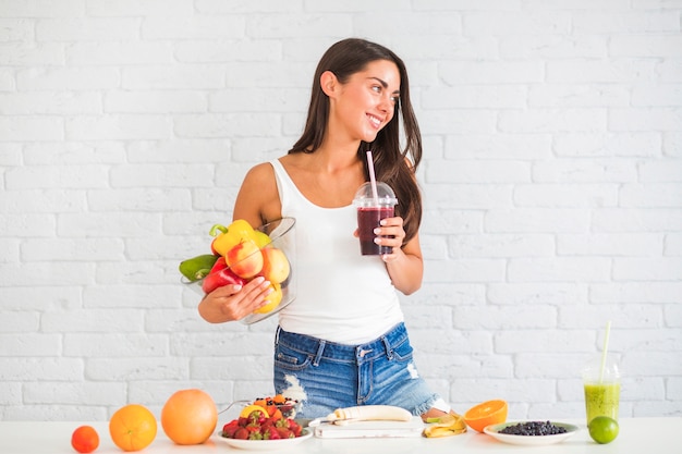 Young woman standing against wall holding bowl of fresh vegetables and fruits and juice