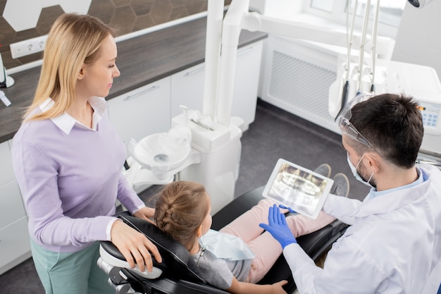 Young woman standing by her little daughter sitting in armchair at the dentist and both looking at x-ray in touchpad