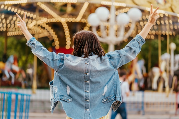 Young woman standing in front of the carrousel with night illumination at the amusement park. Back view