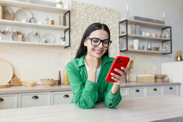 Young woman standing in kitchen at home leaning on table and using mobile phone chatting checking