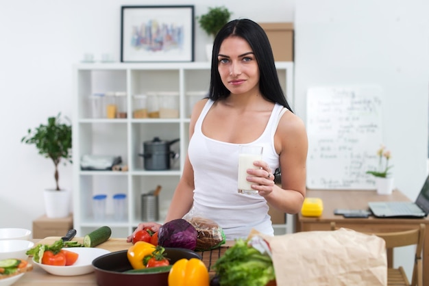 Young woman standing next to kitchen table with fresh vegetables drinking milk in the morning