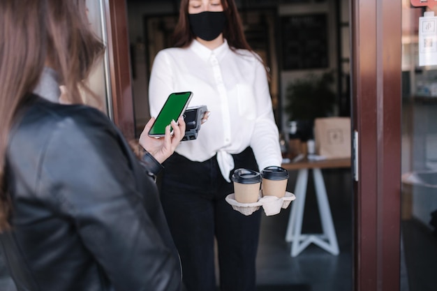 Young woman standing outdoors in mask and paying bill through smartphone using nfc technology in a