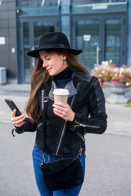 Young woman staying with smartphone and coffee in front of the office.