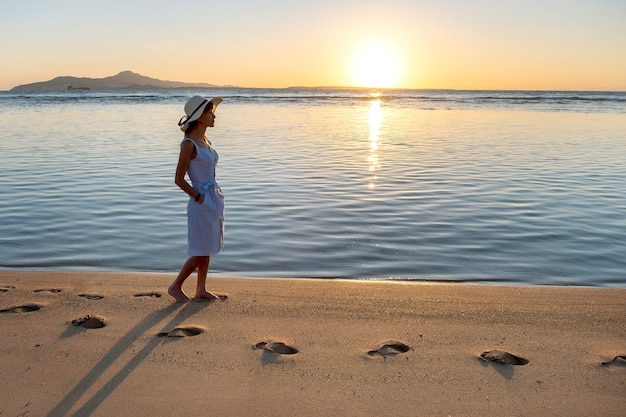 Young woman in straw hat and a dress walking alone on empty sand beach at sunset sea shore. Lonely girl looking at horizon over calm ocean surface on vacation trip.