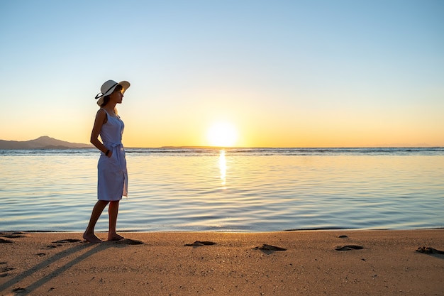 Young woman in straw hat and a dress walking alone on empty sand beach at sunset sea shore