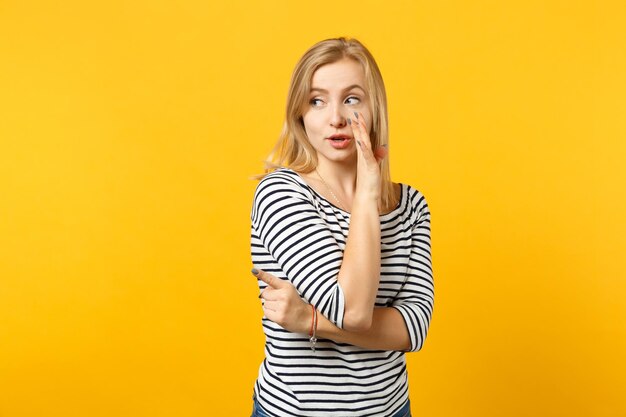 Young woman in striped clothes looking aside, whispers gossip and tells secret with hand gesture isolated on yellow orange background. People sincere emotions, lifestyle concept. Mock up copy space.