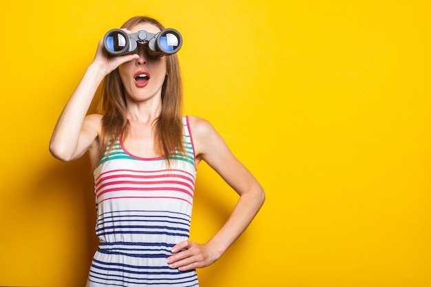 Young woman in a striped dress looks in shock with surprise in binoculars on a yellow space