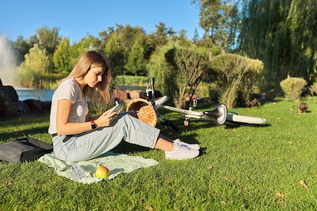 Young woman student 19, 20 years old sitting on grass in park with textbook, girl studies using smartphone, writes in notebook. E-learning, educational technologies, distance learning, self-education