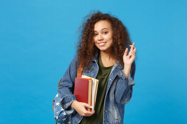 Young woman student in denim clothes and backpack holds books isolated on blue wall