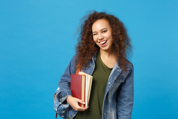 Young woman student in denim clothes and backpack holds books isolated on blue wall