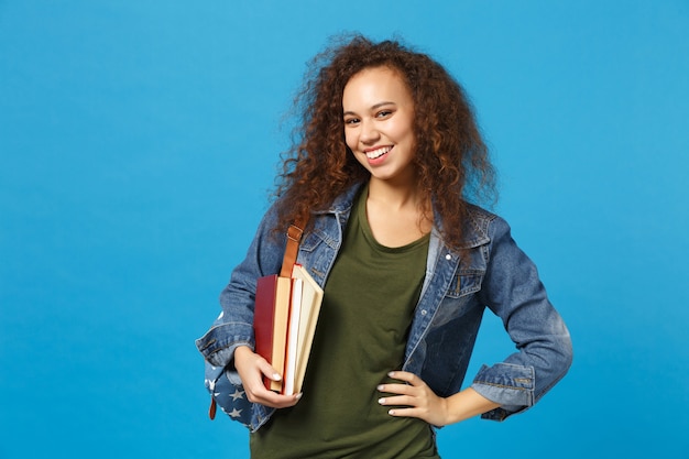 Young woman student in denim clothes and backpack holds books isolated on blue wall