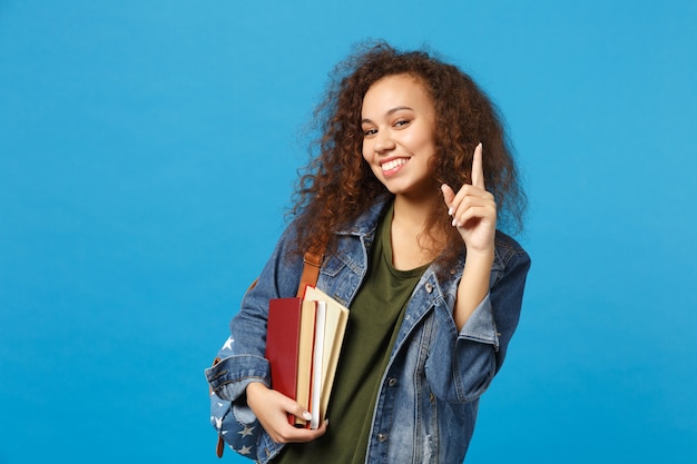 Young woman student in denim clothes and backpack holds books isolated on blue wall