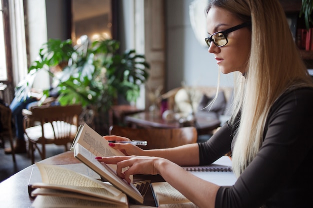 Young woman student of the University. Preparing exam and learning lessons in public library. Sitting at table surrounded by large number of textbooks