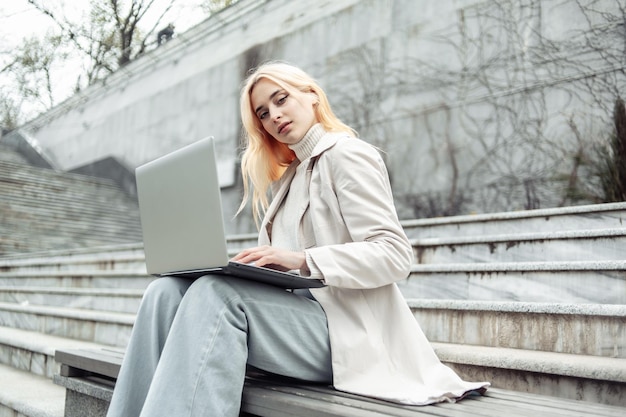 Young woman student uses a laptop while sitting on a bench on the background of the stairs