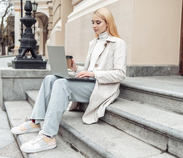 Young woman student uses laptop while sitting on stairs in city