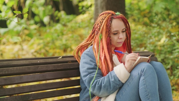 Young woman student with long hair in casual clothes sitting on wooden bench and writing notes in notebook