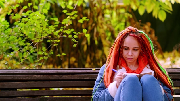 Young woman student with long hair in casual clothes sitting on wooden bench and writing notes in notebook