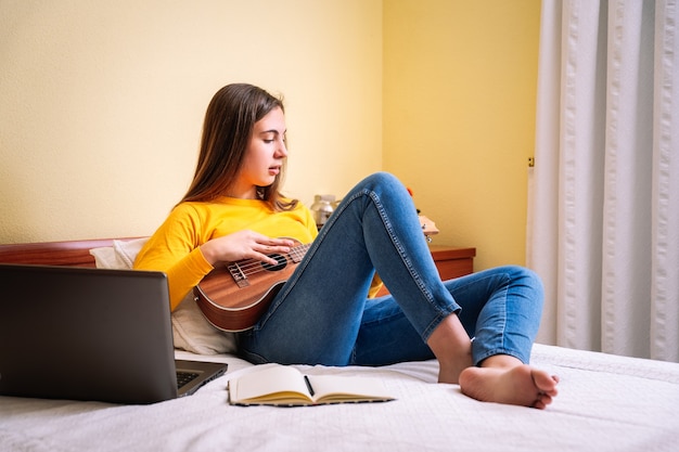 Young woman studied with ukulele on top of bed with laptop