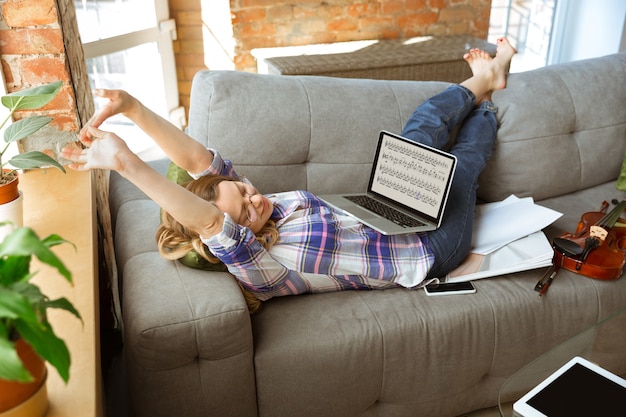 Young woman studying at home during online courses or free information by herself. Becomes musician, violinist while isolated, quarantine against coronavirus spreading. Using laptop, smartphone.