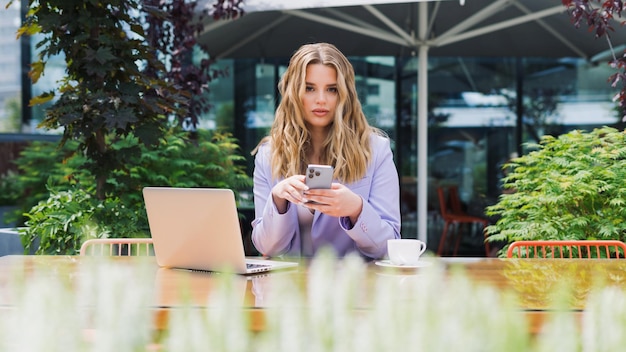 A young woman in a stylish suit is typing a message on her phone while sitting in front of a laptop