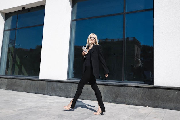 A young woman in a stylish suit with a laptop and a cup of coffee poses against the background of a business center