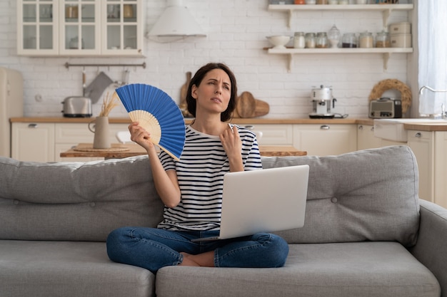 Young woman suffers from heatstroke flat without airconditioner waving fan sitting on couch at home