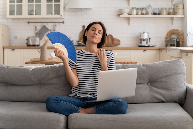 Young woman suffers from heatstroke flat without airconditioner waving fan sitting on couch at home