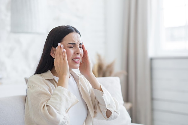 A young woman suffers from a severe headache pressure she is sitting on the couch at home holding