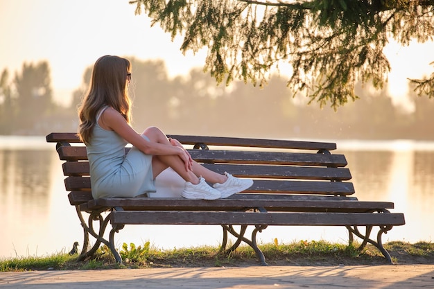 Young woman in summer dress sitting relaxed on lake side bench on warm evening Wellness resting from everyday rush concept