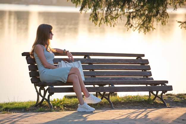 Young woman in summer dress sitting relaxed on lake side bench on warm evening Wellness resting from everyday rush concept