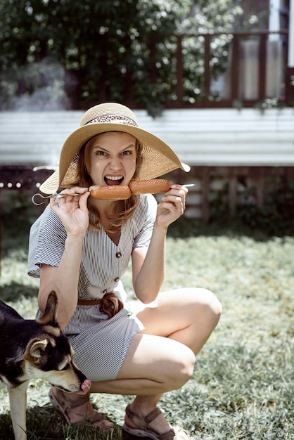 Young woman in summer hat grilling meat outdoors in the backyard, sitting with her dog, giving pet a snack