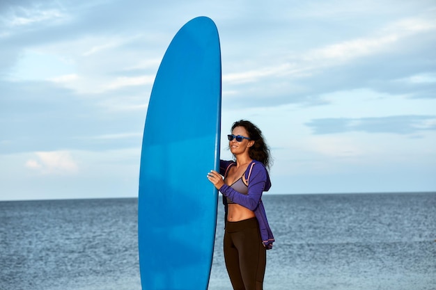 Young woman in sunglasses with surfboard standing on the beach